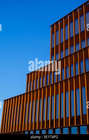 Moderne Bürofassade mit orangefarbenen vertikalen Finnen und reflektierenden blauen Fenstern, geometrischen Wiederholungen und klaren architektonischen Linien unter klarem Himmel. Stockfoto