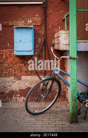 Blaues Fahrrad auf grünem Pfosten neben abblätternder roter Backsteinwand und pastellblauem Briefkasten, urbane Textur und Vintage-Straßenszene, rustikale Farben und abgenutzte Details. Stockfoto
