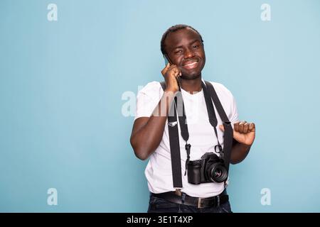 Fröhlicher schwarzer Mann in Hosenträgern spricht auf seinem Handy, stehend in einem Studio mit blauem Hintergrund. Er blickt auf die Kamera mit einem natürlichen und freundlichen Ausdruck, der moderne Kommunikation verkörpert. Stockfoto