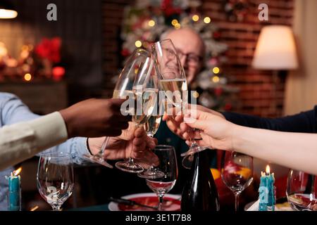 Glückliche Personen, die am weihnachtsabend Gläser anschlagen, Winterferien mit Alkohol und hausgemachten Speisen feiern. Verschiedene Freunde und Familienmitglieder, die am Tisch jubeln, gemütliche Einrichtung. Stockfoto