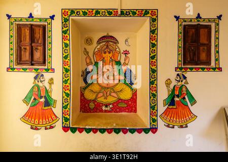 Eine Mauer in Udaipur zeigt ein zentrales Gemälde von Lord Ganesha in einer Nische, flankiert von zwei kleineren Holzfenstern und Figuren von königlichen Wachen. Stockfoto