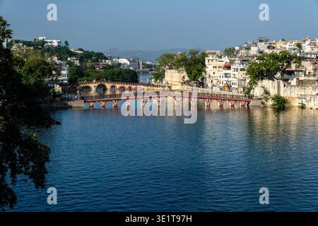 Die Daiji-Fußgängerbrücke überquert das Wasser in Udaipur, mit der gewölbten Chandpole-Brücke und Hügeln, die in der Ferne hinter der städtischen Skyline sichtbar sind. Stockfoto