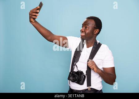 Junger schwarzer Mann, der ein Selfie mit seinem Handy macht. afroamerikanischer männlicher Mann mit einer Kamera um den Hals greift sein Strapsband, während er ein Video auf seinem Mobilgerät aufnimmt. Stockfoto