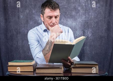 Ein Mann, der in das Lesen eines Buches eingetaucht ist. Der Mann ist vierzig Jahre alt, trägt ein weiß-blaues Hemd und sitzt an einem Schreibtisch, auf dem andere Bücher stehen Stockfoto