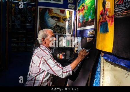 In einem Atelier in Udaipur sitzt ein Künstler im Kreuz, während er einen weißen Stier auf ein schwarzes Kleid malt, umgeben von verschiedenen farbenfrohen Kunstwerken. Stockfoto