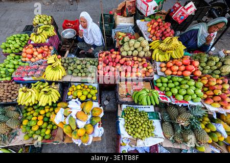 Marktszene in Udaipur mit zwei Frauen, die hinter einer großen Ausstellung tropischer Früchte wie Bananen, Äpfel und Papayas sitzen. Stockfoto