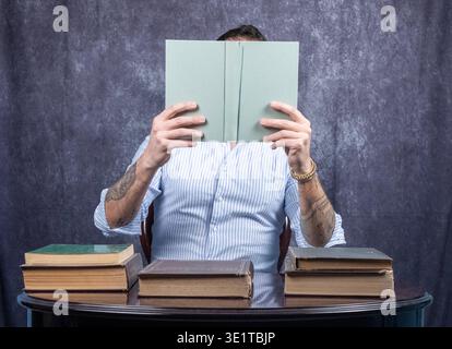 Mann mit Gesicht, bedeckt von dem Buch, das er liest. Der Mann trägt ein weiß-blaues Hemd und sitzt an einem Schreibtisch mit anderen Büchern darauf Stockfoto