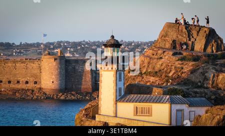 Blick aus der Vogelperspektive auf den weißen Leuchtturm von Ile Louet vor den zerklüfteten, sonnendurchfluteten Felsen, wo sich die Figuren versammeln, im Kontrast zum ruhigen blauen Meer, P Stockfoto