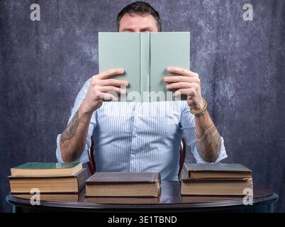 Mann mit Gesicht, bedeckt von dem Buch, das er liest. Der Mann trägt ein weiß-blaues Hemd und sitzt an einem Schreibtisch mit anderen Büchern darauf Stockfoto