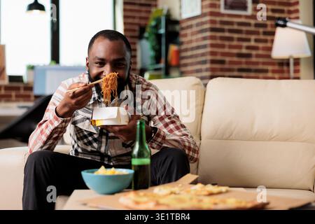 afroamerikanischer Mann, der Essstäbchen zum Mitnehmen von Nudeln zu Hause verwendet und sich einen Film auf dem fernsehsender ansieht. Essen Sie eine Mahlzeit zum Mitnehmen aus der Box und genießen Sie Filme im Fernsehen. Stockfoto