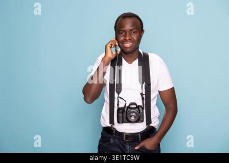 Ein lächelnder schwarzer Mann, der mit einer Hand in der Tasche in die Kamera schaut, während er einen Anruf auf seinem Mobilgerät entgegennimmt. Ein Fotograf, der Hosenträger trägt, posiert für ein Studio-Shooting mit einem Telefon am Ohr. Stockfoto