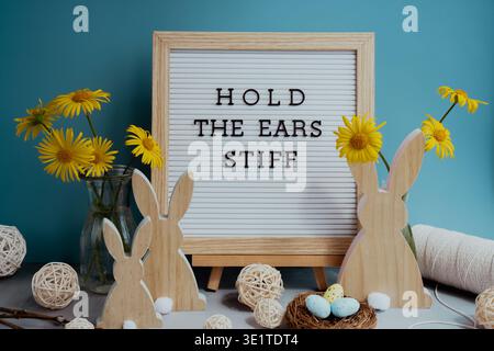 HALTEN SIE DIE OHREN STEIF Text auf der Brieftafel mit Hasen Holzfiguren Eier und frische Blumen. Osterkompositionsinhalt Stillleben. Christliche Symbole der Frühlingsferien Stockfoto