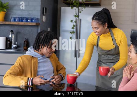 Afroamerikanischer Server in Schürze serviert rote Tassen auf einem Hochglanztisch, während Kunden das Telefon halten Stockfoto