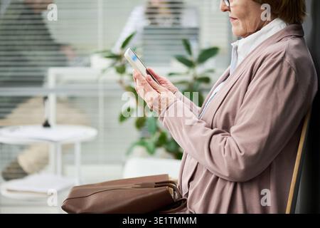 Ältere Frau, die ihr Smartphone benutzt, während sie drinnen in der Nähe des Fensters sitzt Stockfoto