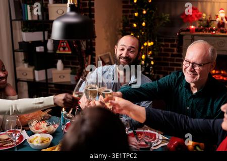 Vielfältige Menschen zwitschern Weingläser am weihnachtsabend-Dinner, feiern Feiertage mit Alkohol und hausgemachten Speisen. Freunde und Familie jubeln mit Getränken am Tisch, gemütliche Dekoration. Stockfoto