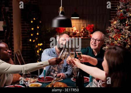 Verschiedene Personen zwitschern beim Abendessen am heiligabend die Alkoholgläser, feiern die Feiertagstradition mit Wein und hausgemachtem Essen. Freunde und Familie jubeln mit Drinks am Tisch, gemütlichen Ornamenten. Stockfoto