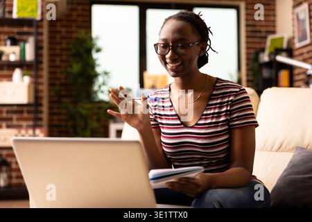 Lächelnde schwarze Frau mit Stift in der Hand, winkt auf dem Bildschirm des Geräts, nimmt von zu Hause aus an einem Online-Business-Meeting Teil. afroamerikanische Unternehmerin, die sich über Videoanrufe mit Teammitgliedern verbindet. Stockfoto