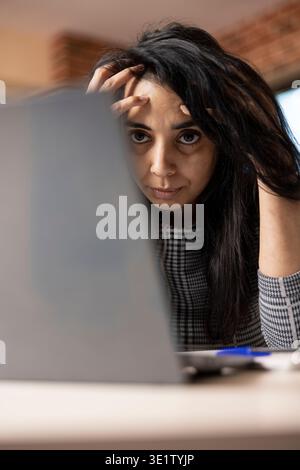 Überarbeiteter weiblicher Profi, der mit Händen auf dem Kopf auf den Laptop starrt und sich über die Arbeitsbelastung ärgert. Frustrierte indianerin, die von zu Hause aus arbeitet und geistige Erschöpfung erlebt. Stockfoto