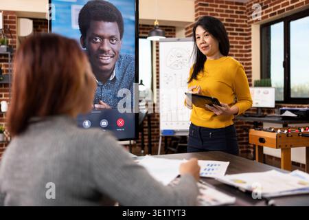 Asiatische Frauen führen ein virtuelles Treffen mit Kunden und präsentieren strategische Erkenntnisse. Die Beraterin präsentiert Projektideen mit einem Tablet, während die Mitarbeiterin am Schreibtisch sitzt und mit einem Mann in einem Videoanruf Pläne teilt. Stockfoto