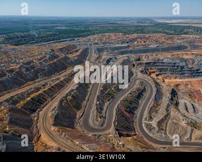 Aus der Vogelperspektive führt eine kurvenreiche Straße durch die schroffe, geschichtete Landschaft des riesigen Eisenerzsteinbruchs, im Kontrast zu den fernen grünen Wäldern Kryvy Stockfoto