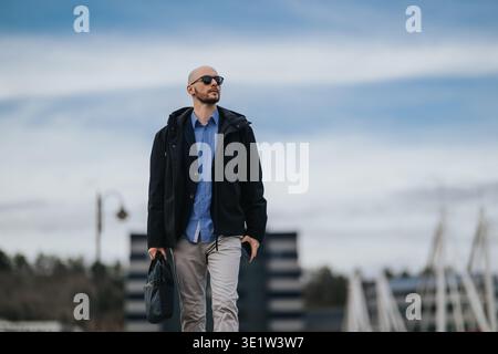 Geschäftsmann, der mit Tasche und Sonnenbrille im urbanen Hafenviertel draußen spaziert Stockfoto