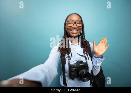 Die lächelnde Fotografin macht eine Hello-Geste und nimmt Videoinhalte mit einer dslr um den Hals auf. Afroamerikanerin winkt ihrem Online-Publikum zu und teilt Updates über ihre Fotografie-Reise. Stockfoto