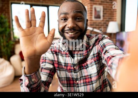 Lächelnder Geschäftsmann, der das Handy für Videoanrufe benutzt und in freundlicher Begrüßung zur Kamera winkt. Glücklicher männlicher Freelancer sitzt im Wohnzimmer und macht eine Hello-Hand-Geste während einer fröhlichen Online-Konversation. Stockfoto