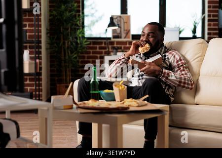 Männliche Person, die zu Hause Fast-Food-Delivery-Mahlzeit isst, Filme im Fernsehen ansieht und Freizeit genießt. Es werden Burger vom Take-out-Paket, Chips und Bierflaschen im tv-Film serviert. Stockfoto