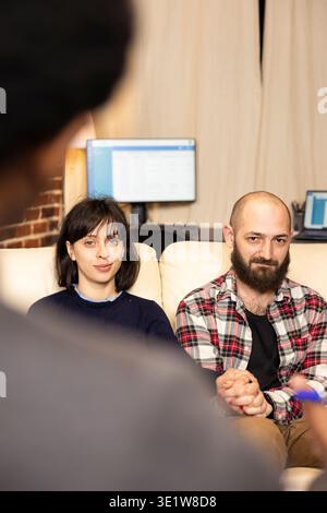 Ein lächelndes Paar im Büro des Psychologen sitzt zusammen auf der Couch, hält die Hände und blickt mit ruhigem Ausdruck in die Kamera. Ehemann und Ehefrau zeigen nach erfolgreicher Therapiesitzung Positivität und Erleichterung. Stockfoto