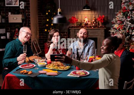 Frau, die Freunde nimmt weihnachtsfeier Gruppenfoto auf Handy, verschiedene Menschen Porträt auf festlichen Esstisch. Große Familientreffen, Weihnachten feiern, traditionelles Essen essen, trinken Stockfoto