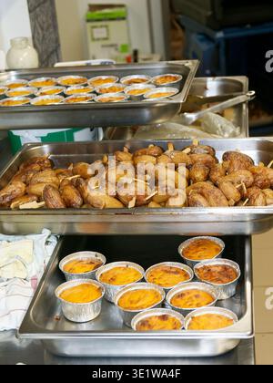 Mehrere Tabletts mit zubereiteten Speisen, einschließlich Bratkartoffeln und Backgerichten, in einer professionellen Küche für Catering-Service angeordnet. Szene-Highlights Stockfoto