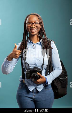 Optimistische schwarze Frau, die eine dslr-Kamera hält und eine Geste zur Genehmigung macht, bereit für eine Kundensitzung. afroamerikanische Fotografin, die die Daumen hoch hält, steht vor blauem Hintergrund. Stockfoto