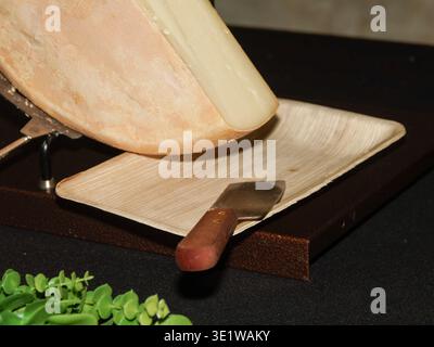 Nahaufnahme eines Raclettekäses, der von einem Rad geschnitten und mit einem Käsemesser auf einem Teller serviert wird. Traditionelles alpenländisches Gericht mit geschmolzenem Käse Stockfoto