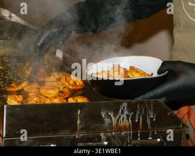 Nahaufnahme gegrillter Garnelen, die auf einem heißen Grill gekocht werden, wobei ein Koch schwarze Handschuhe trägt und frisch gegrillte Meeresfrüchte in einer Schüssel serviert. Stockfoto