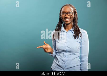 Die schwarze Frau zeigt nach links und lenkt den Fokus auf leere Räume, perfekt für Werbung. Die glückliche afroamerikanische Dame in blauem Hemd bewegt sich seitlich mit dem Zeigefinger und weist auf den Bereich der Produktplatzierung hin. Stockfoto