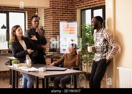 Eine Gruppe multiethnischer Kollegen lacht zusammen und macht eine kurze Pause in einem professionellen Arbeitsplatz. Ein vielfältiges Startup-Team, das nach einem langen Arbeitstag abhängt und Kaffee getrunken hat. Stockfoto