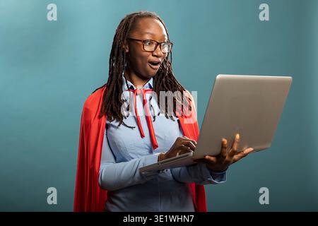 Überraschte afroamerikanerin mit rotem cape und blauem Hemd, die auf den Laptop-Bildschirm blickt. Glückliche schwarze Frau mit schockierter Reaktion, drückt Begeisterung über gute Nachrichten aus. Stockfoto