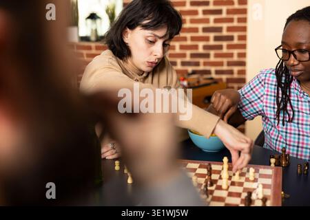 Fokussierte weiße Frau, die sich während des lässigen Spiels in Schach bewegt. Kaukasische Frau macht sich auf den Weg, mit Getränken und Snacks auf dem Tisch, während Freunde das freundliche Spiel im gemauerten Raum genießen. Stockfoto