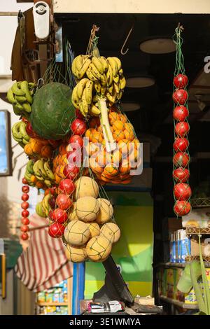 Obststand mit hängenden Netztüten gefüllt mit Bananen, Orangen, Melonen, Tomaten und einer großen grünen Frucht, die ein farbenfrohes Arrangement schaffen Stockfoto