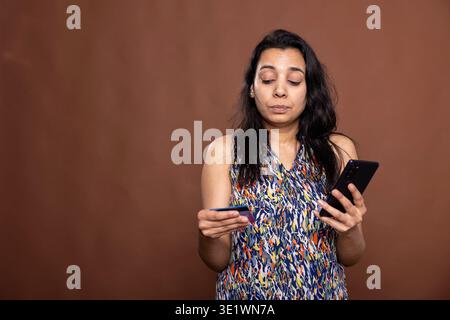 Selbstbewusste weibliche Person, die ein Mobilgerät nutzt, um Online-Transaktionen im Studio mit braunem Hintergrund abzuschließen. Indische Frau mit Smartphone und Debitkarte, die sichere digitale Zahlung durchführt. Stockfoto
