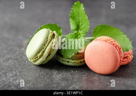 Französische rosafarbene und grüne Macarons Kekse mit Minzblättern auf dunklem Steinhintergrund. Himbeer- und Pistazienmakronen aus der Nähe. Stockfoto