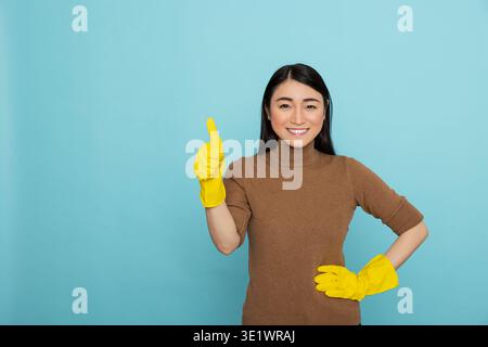 Aufgeregte asiatische Haushälterin, die gelbe Gummihandschuhe trägt, die Daumen hoch hält und selbstbewusst vor isoliertem Hintergrund lächelt. Die Reinigungsfrau zeigt eine Geste der Zustimmung und ist mit den abgeschlossenen Reinigungsaufgaben zufrieden Stockfoto