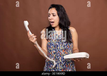 Wütende hübsche Frau, die während eines intensiven Anrufs in den Festnetzempfänger ruft. Indische Frau, die das Telefon hält und ein hitziges Gespräch im Studio mit braunem Hintergrund führt. Stockfoto