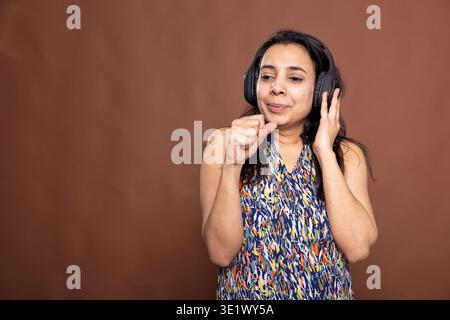 Junge Erwachsene, die schnurlose Kopfhörer trägt und ihre Faust zum Mund hebt, als würde sie mitsingen. Indisches Modell, das vollständig in die Musik eingetaucht ist, das Audioerlebnis genießt und ihre Hand wie ein Mikrofon verwendet. Stockfoto