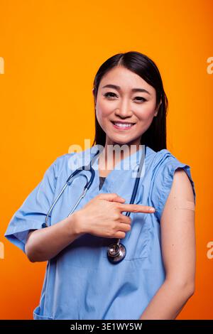 Asiatische Krankenschwester in medizinischer Uniform, zeigt ihren geimpften Arm und lächelt in die Kamera. Eine junge Frau mit blauen Peelings und einem Stethoskop um den Hals zeigt auf die Bandage. Stockfoto