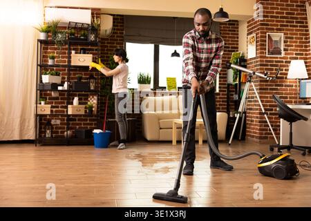 Verschiedene romantische Partner, die gemeinsam in der Wohnung wohnen. Kaukasische Ehefrau wischt Staub auf den Regalen ab, während schwarzer Ehemann Staubsauger verwendet, wodurch ein ordentliches und hygienisches Zuhause mit Teamarbeit geschaffen wird. Stockfoto
