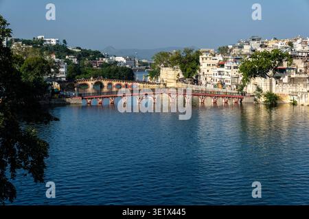 Die Daiji-Fußgängerbrücke überquert das Wasser in Udaipur, mit der gewölbten Chandpole-Brücke und Hügeln, die in der Ferne hinter der städtischen Skyline sichtbar sind. Udaipu Stockfoto