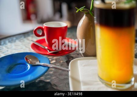 Orangener Espresso in hohem Glas, rote Tasse und blaue Teller auf Glastisch im Café. Stockfoto