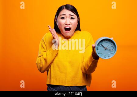 Asiatische Frau in gelbem Pullover hält eine blaue Uhr mit schockiertem Ausdruck, die die Dringlichkeit und den Stress des Zeitmanagements zum Ausdruck bringt. Weibliche Person, die überrascht und besorgt aussah und sich spät für einen Termin verspätet. Stockfoto