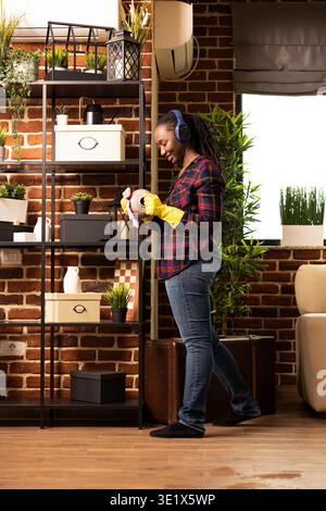Lächelnde afroamerikanische Hausfrau steht in der Nähe von Regalen und putzt die Vase mit einem Tuch, während sie Musik hört. Schwarze Frau, die Kopfhörer und Handschuhe trägt, macht gerne Hausarbeiten in gemauertem Haus. Stockfoto